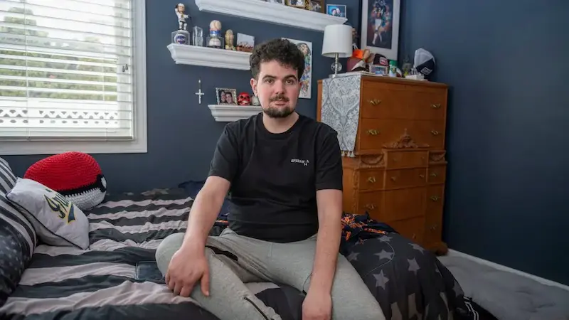 A young man with dark, curly hair and a beard sits on a striped bed in a bedroom with dark blue walls, looking at the viewer.